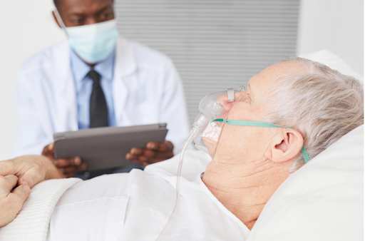A senior man wearing an oxygen mask, assisted by a healthcare worker in a clinical setting.